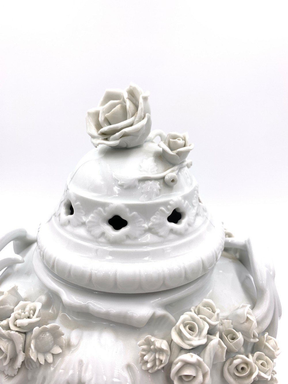 A white glazed porcelain urn featuring applied floral garlands, a pedestal base, and a pierced lid topped with a rose finial. View VI