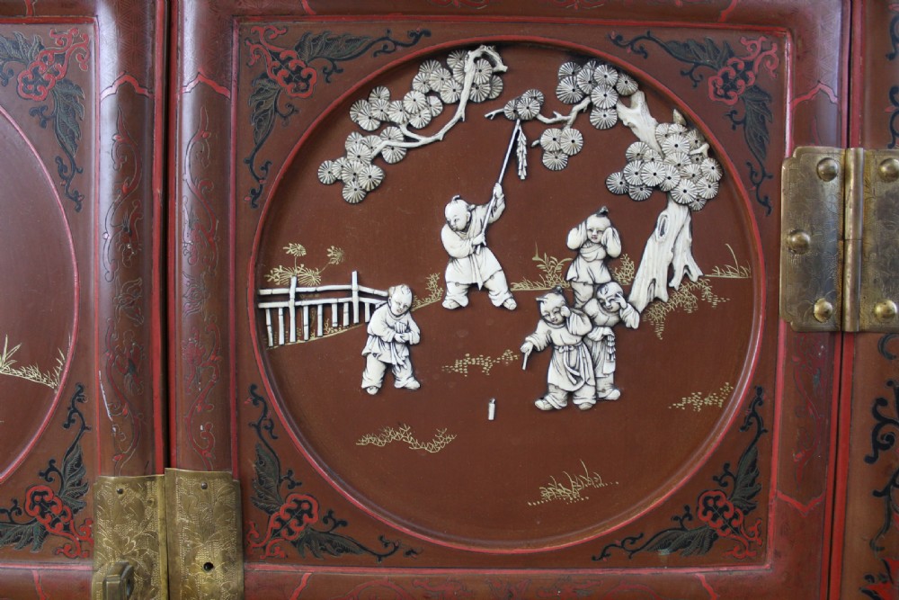 A 19th-century Chinese red lacquer cabinet featuring two doors decorated with carved white relief panels depicting figures, birds, and botanical motifs, fitted with brass lockplates. View IX