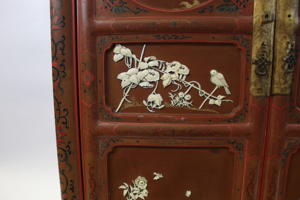 A 19th-century Chinese red lacquer cabinet featuring two doors decorated with carved white relief panels depicting figures, birds, and botanical motifs, fitted with brass lockplates. View X
