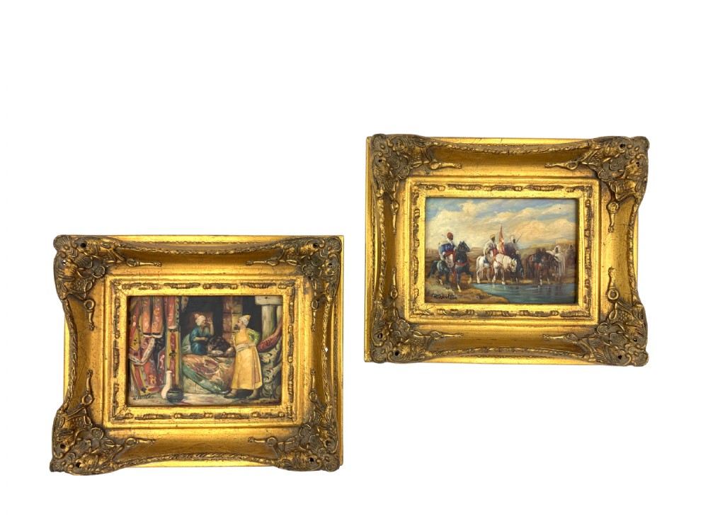 A pair of framed Orientalist oil paintings on board in heavily ornamented gold gilt frames, depicting Middle Eastern figures indoors and riders watering horses outdoors.