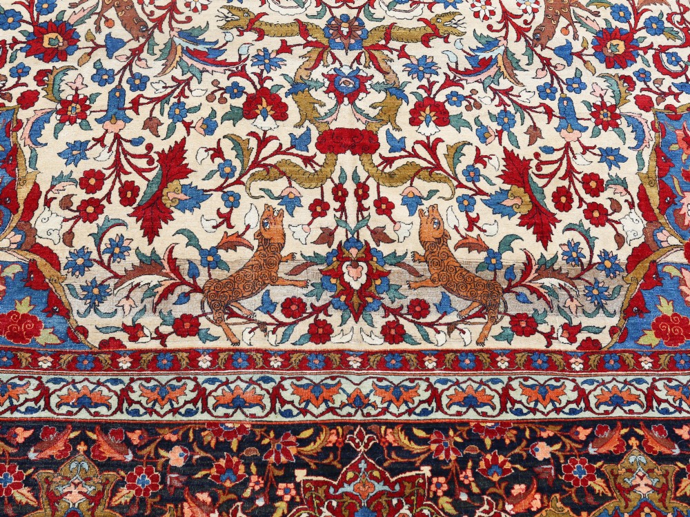 An antique Persian Isfahan wool carpet featuring a central red and blue medallion on an ivory field surrounded by detailed floral and animal patterns with a dark blue border. View II