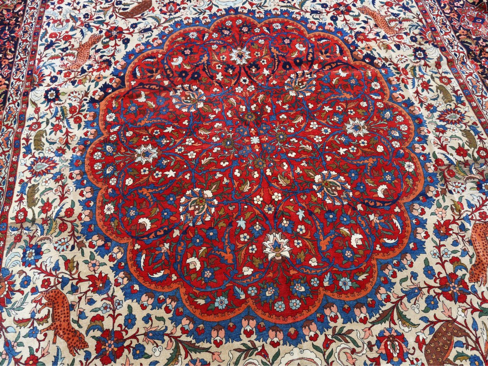 An antique Persian Isfahan wool carpet featuring a central red and blue medallion on an ivory field surrounded by detailed floral and animal patterns with a dark blue border. View III