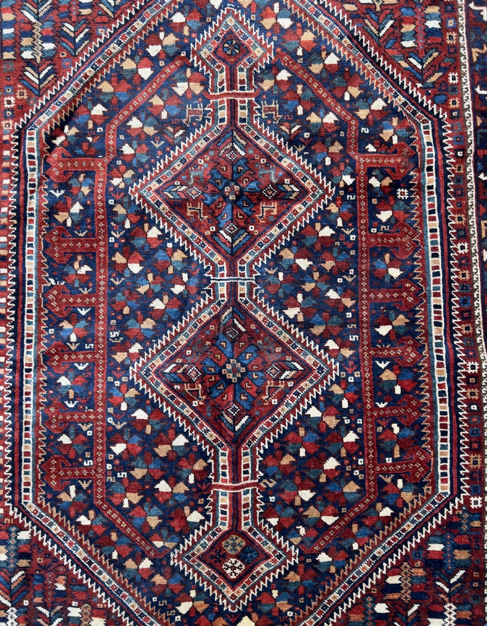An antique hand-knotted wool Qashqai rug featuring geometric diamond medallions surrounded by dense stylized motifs in shades of red, navy blue, and cream with fringed ends. View II
