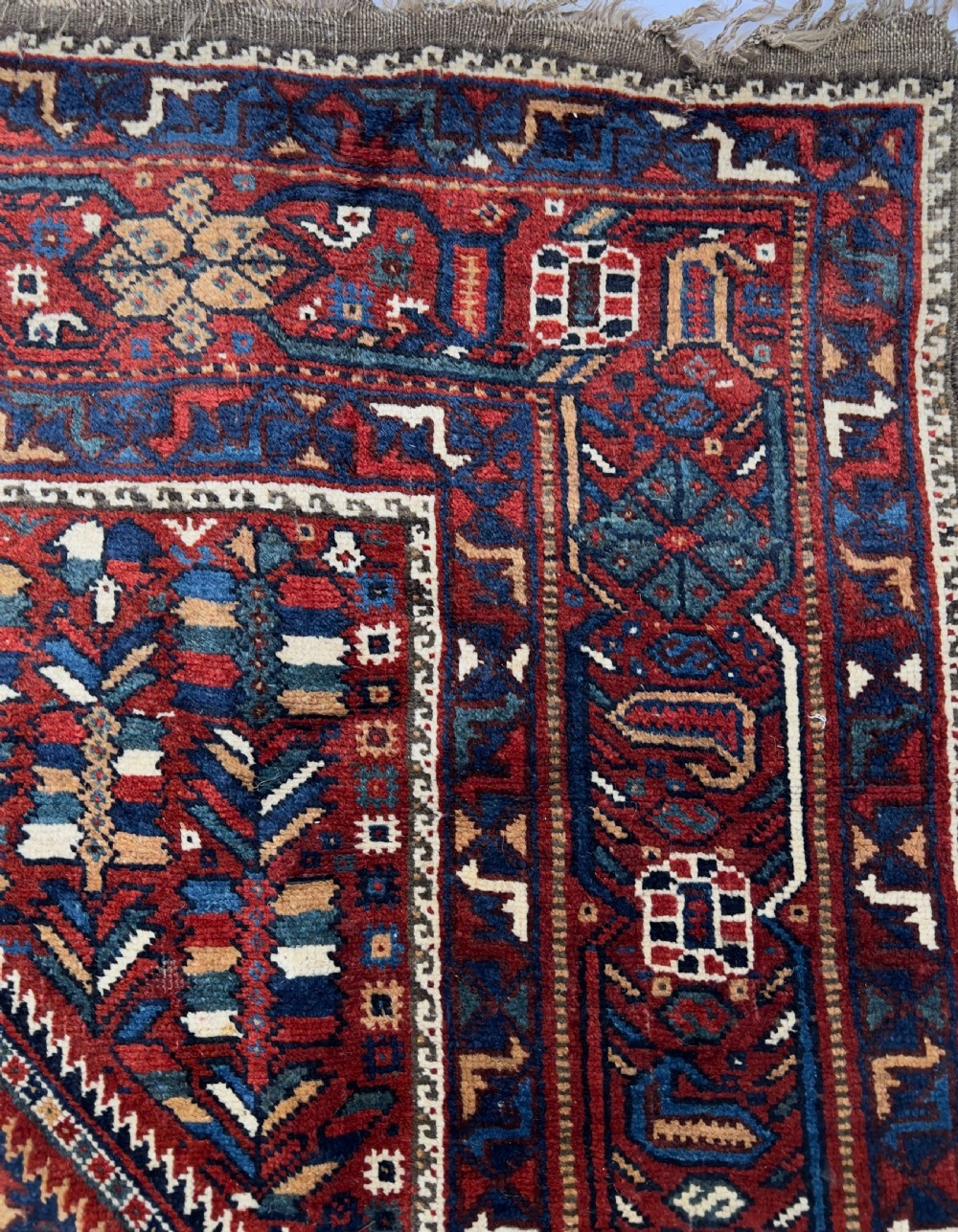 An antique hand-knotted wool Qashqai rug featuring geometric diamond medallions surrounded by dense stylized motifs in shades of red, navy blue, and cream with fringed ends. View III