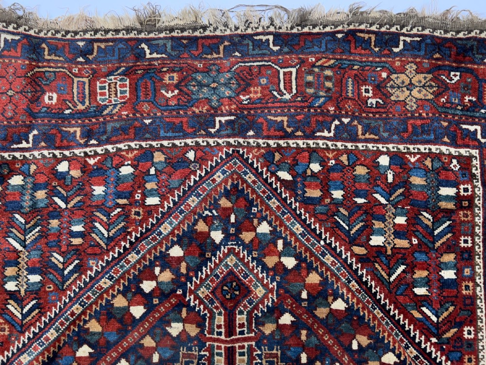 An antique hand-knotted wool Qashqai rug featuring geometric diamond medallions surrounded by dense stylized motifs in shades of red, navy blue, and cream with fringed ends. View IV