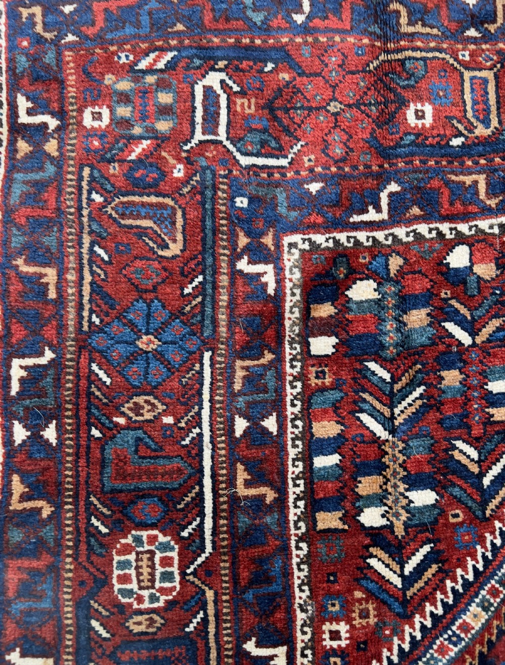 An antique hand-knotted wool Qashqai rug featuring geometric diamond medallions surrounded by dense stylized motifs in shades of red, navy blue, and cream with fringed ends. View V