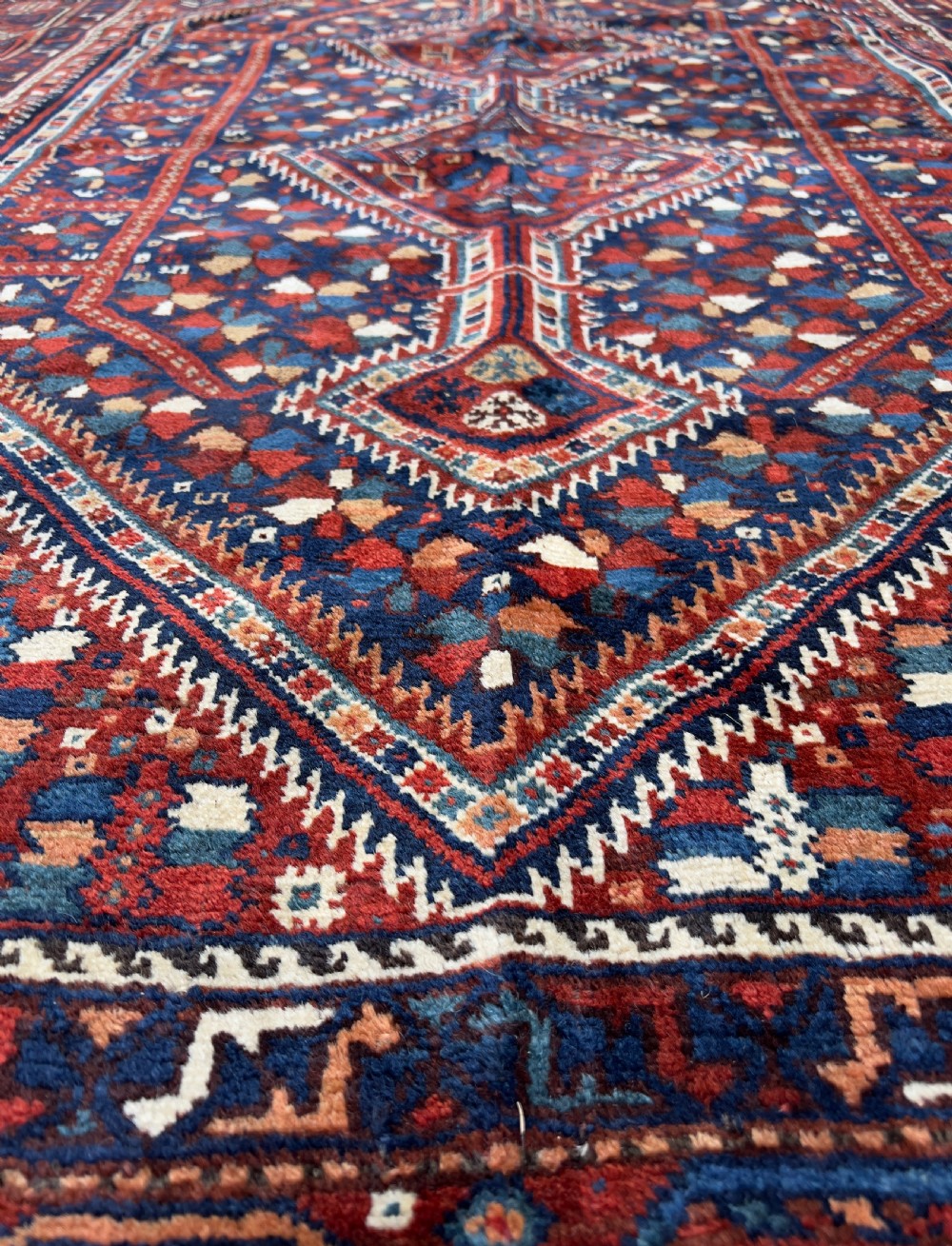 An antique hand-knotted wool Qashqai rug featuring geometric diamond medallions surrounded by dense stylized motifs in shades of red, navy blue, and cream with fringed ends. View VI