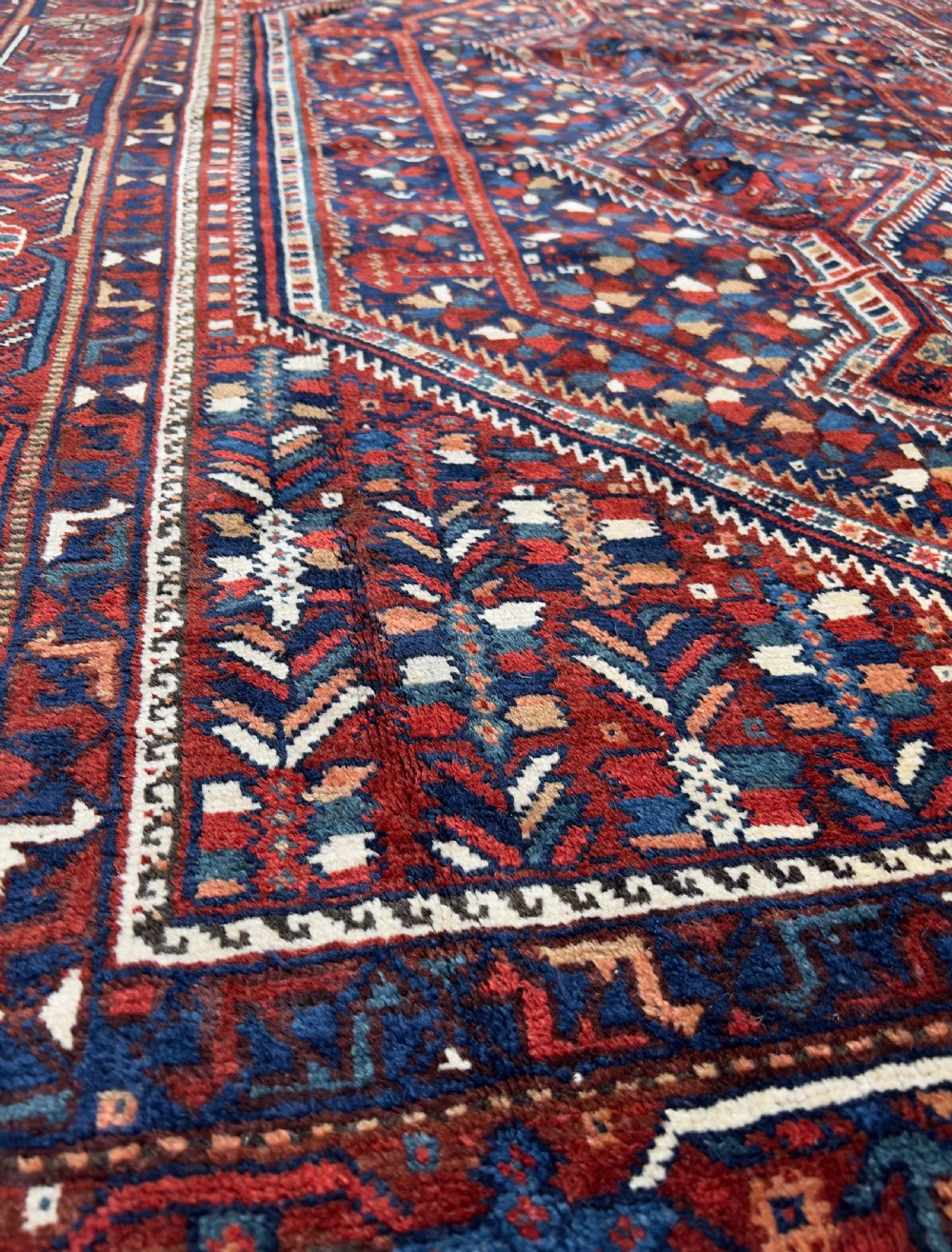An antique hand-knotted wool Qashqai rug featuring geometric diamond medallions surrounded by dense stylized motifs in shades of red, navy blue, and cream with fringed ends. View VII