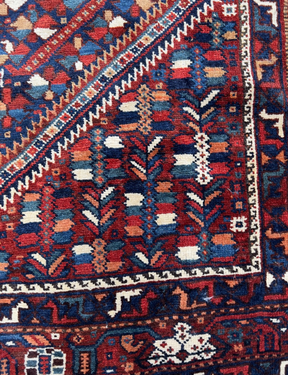 An antique hand-knotted wool Qashqai rug featuring geometric diamond medallions surrounded by dense stylized motifs in shades of red, navy blue, and cream with fringed ends. View VIII