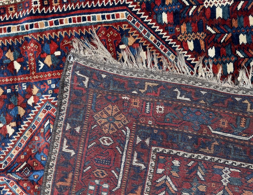 An antique hand-knotted wool Qashqai rug featuring geometric diamond medallions surrounded by dense stylized motifs in shades of red, navy blue, and cream with fringed ends. View IX