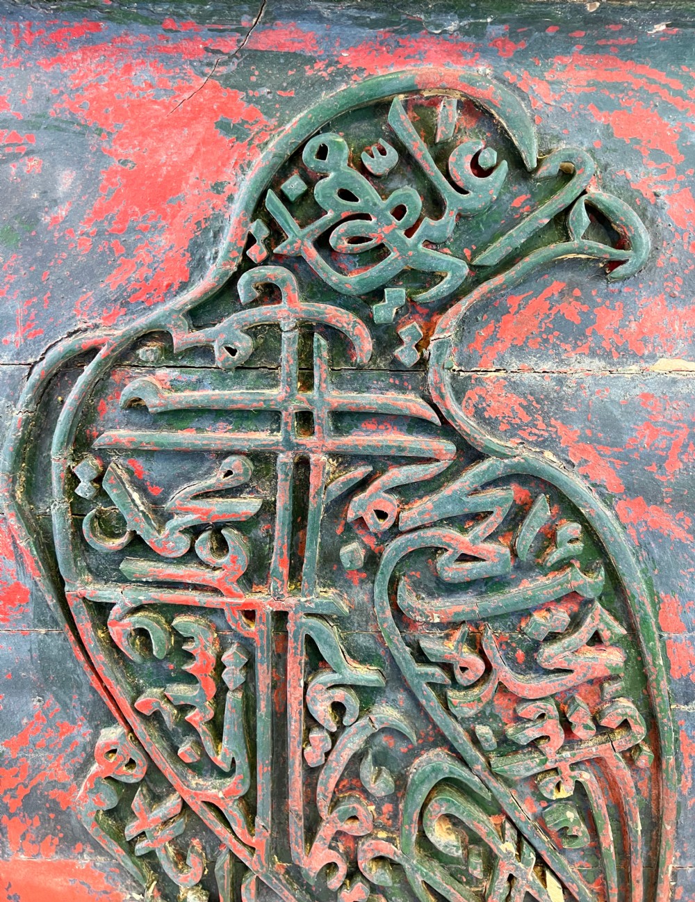 A rectangular carved wooden panel featuring a falcon rendered in green Arabic calligraphy against a distressed red background, surrounded by a carved floral border. View II