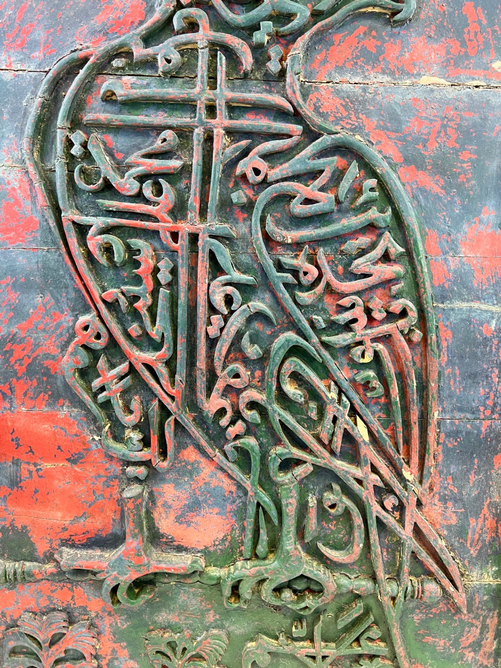 A rectangular carved wooden panel featuring a falcon rendered in green Arabic calligraphy against a distressed red background, surrounded by a carved floral border. View IV