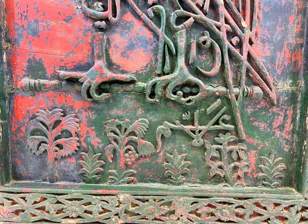 A rectangular carved wooden panel featuring a falcon rendered in green Arabic calligraphy against a distressed red background, surrounded by a carved floral border. View V