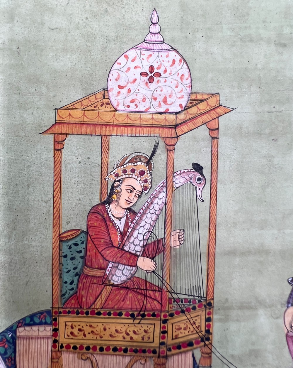 An Indian miniature painting on paper depicting a woman playing a harp within a canopy atop a composite camel, set against a pale green field and framed by a dark floral border. View V