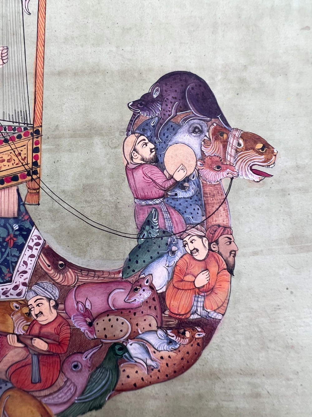 An Indian miniature painting on paper depicting a woman playing a harp within a canopy atop a composite camel, set against a pale green field and framed by a dark floral border. View VI
