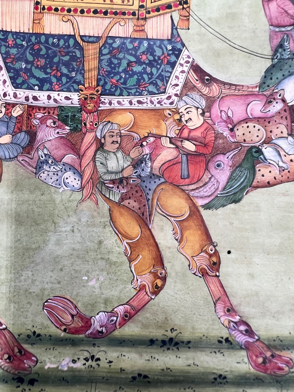 An Indian miniature painting on paper depicting a woman playing a harp within a canopy atop a composite camel, set against a pale green field and framed by a dark floral border. View VII