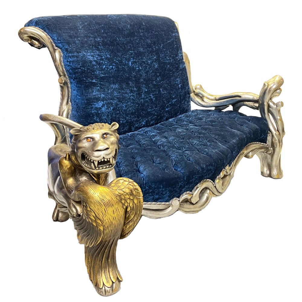 An ornate two-seater sofa featuring deep blue crushed velvet upholstery and a gilded wooden frame carved with a lion on one armrest and branch motifs on the other.