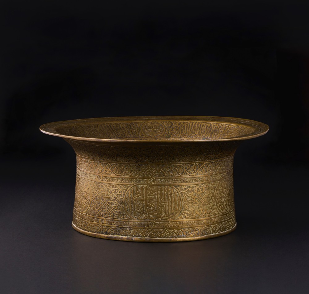 A 16th-century Mamluk brass basin featuring a wide flared rim, intricate hand-engraved Arabic calligraphy, arabesques, and geometric patterns.