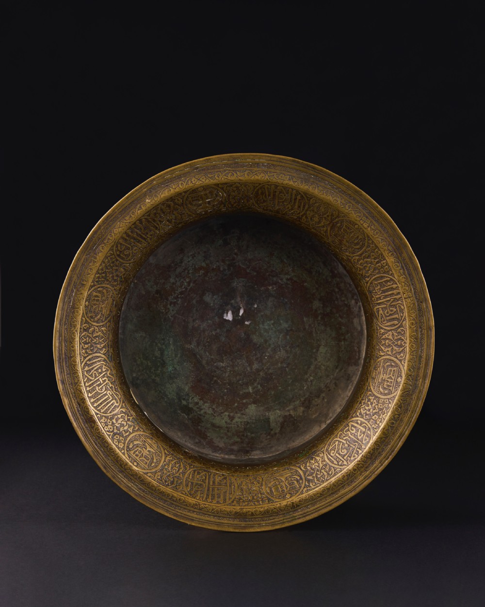 A 16th-century Mamluk brass basin featuring a wide flared rim, intricate hand-engraved Arabic calligraphy, arabesques, and geometric patterns. View V