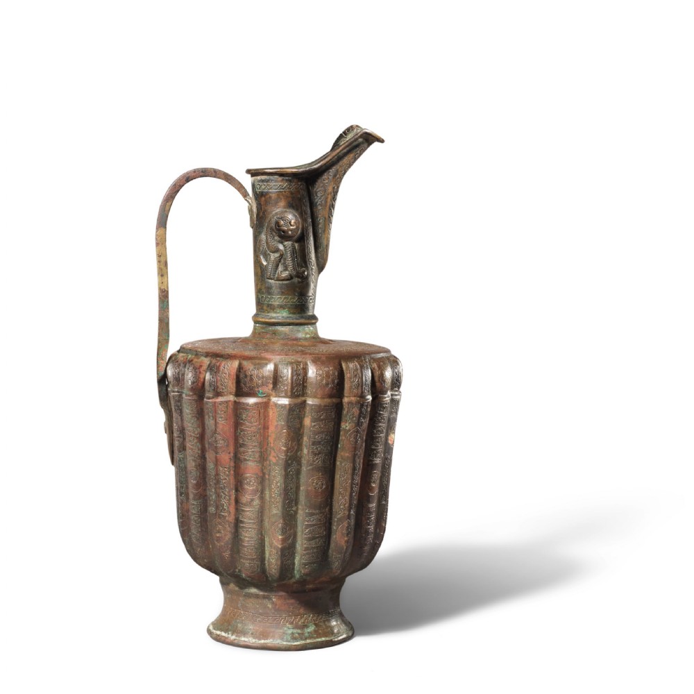 A 12th-century Persian bronze ewer featuring a fluted body, high handle with lion motif, raised spout, and copper and silver inlaid calligraphic bands. View V