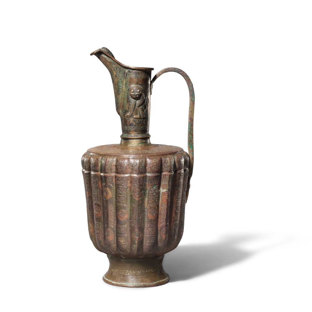 A 12th-century Persian bronze ewer featuring a fluted body, high handle with lion motif, raised spout, and copper and silver inlaid calligraphic bands.