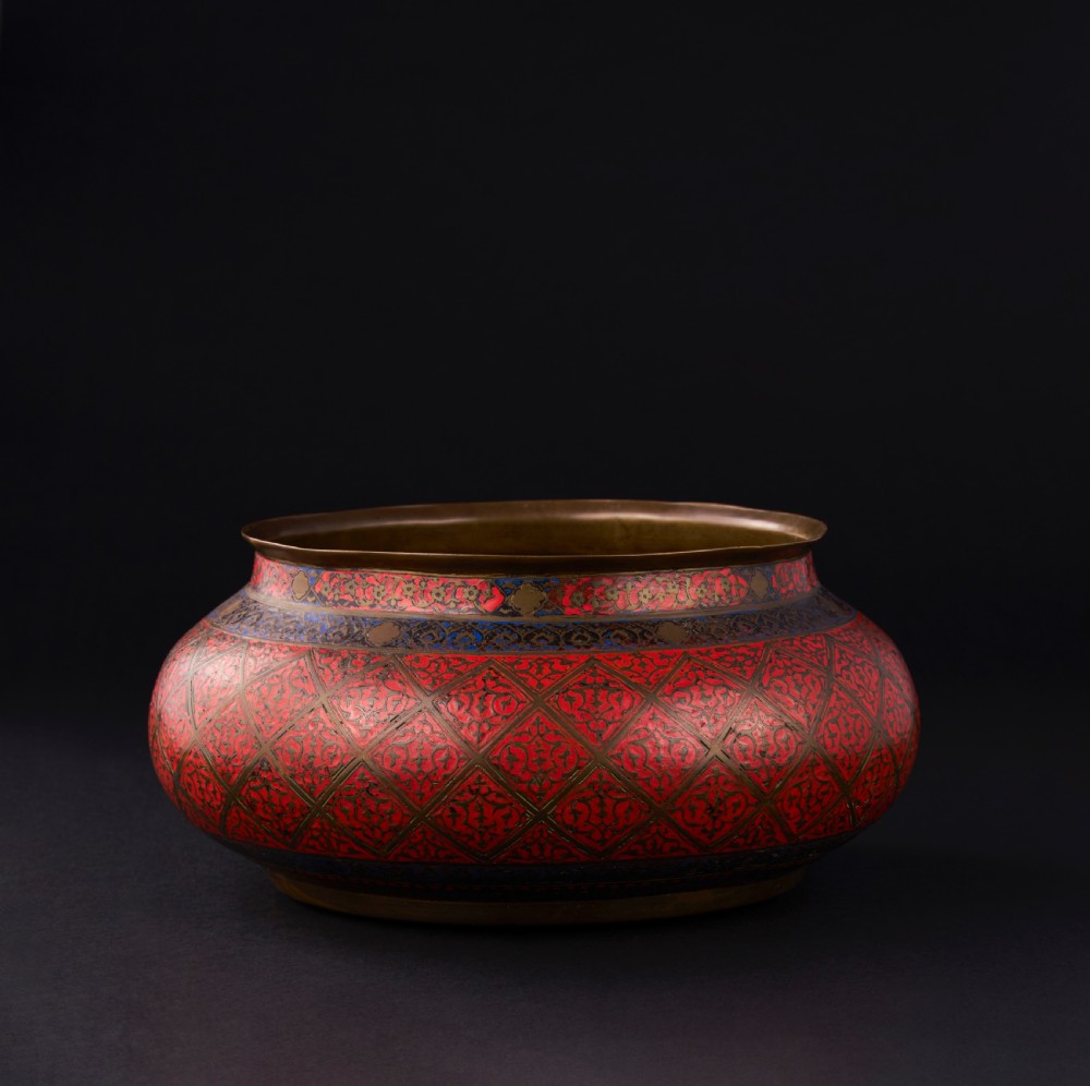 A bulbous Safavid period brass bowl featuring red and blue enamelled geometric lattice patterns around its body.