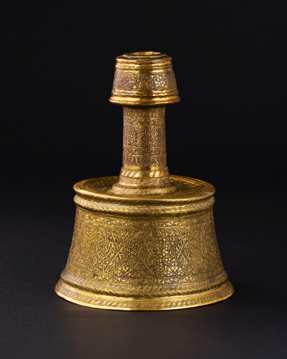 A 15th-century Mamluk brass candlestick featuring a wide flared base and cylindrical stem decorated with intricate geometric and arabesque engravings.
