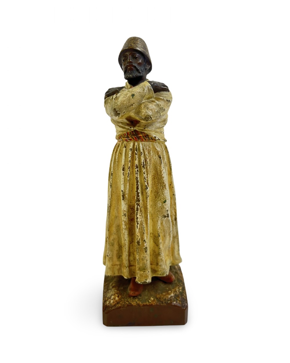 A cold painted bronze statuette depicting a standing whirling dervish with arms crossed, wearing a tall hat and long cream robe.