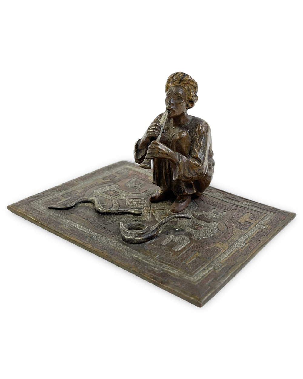 A cold-painted bronze sculpture of a seated snake charmer playing a flute on a rectangular rug base with two coiled snakes.