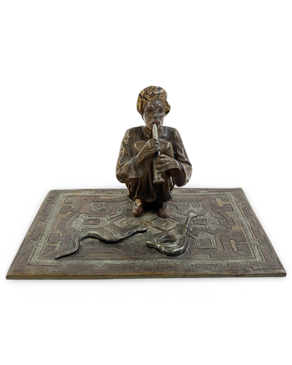 A cold-painted bronze sculpture of a seated snake charmer playing a flute on a rectangular rug base with two coiled snakes. View II