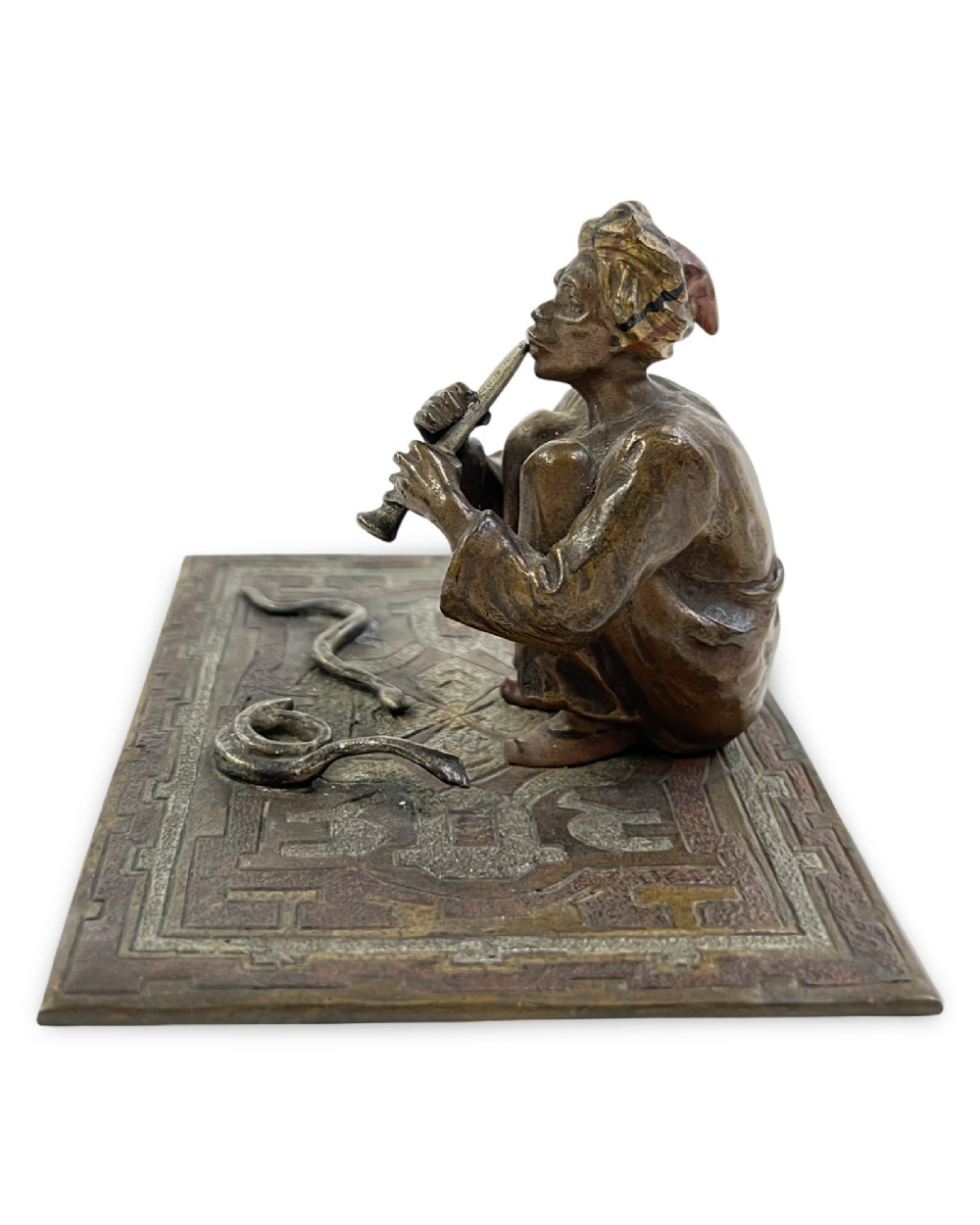 A cold-painted bronze sculpture of a seated snake charmer playing a flute on a rectangular rug base with two coiled snakes. View III