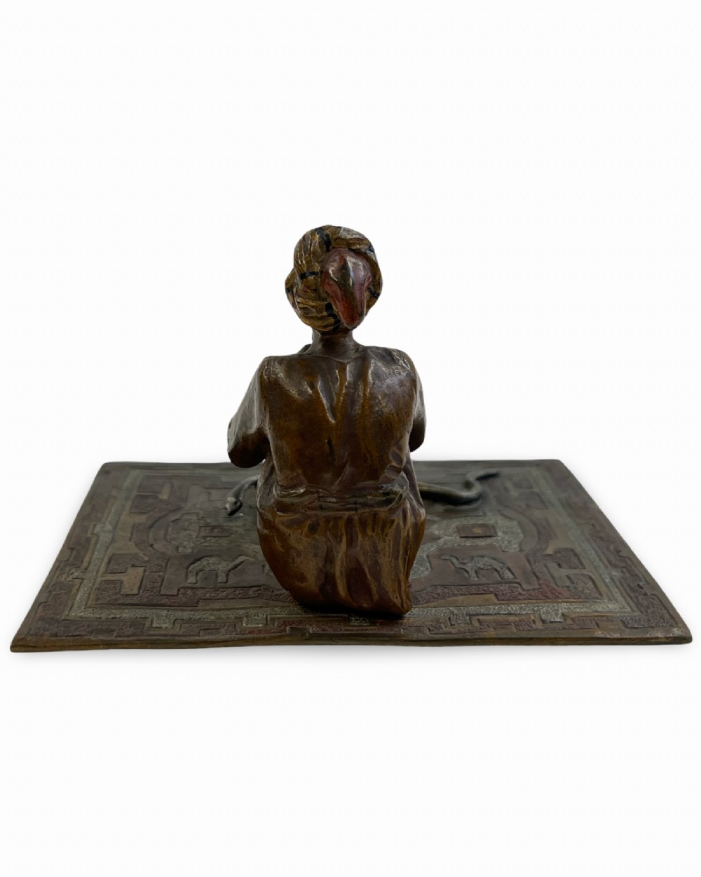 A cold-painted bronze sculpture of a seated snake charmer playing a flute on a rectangular rug base with two coiled snakes. View IV