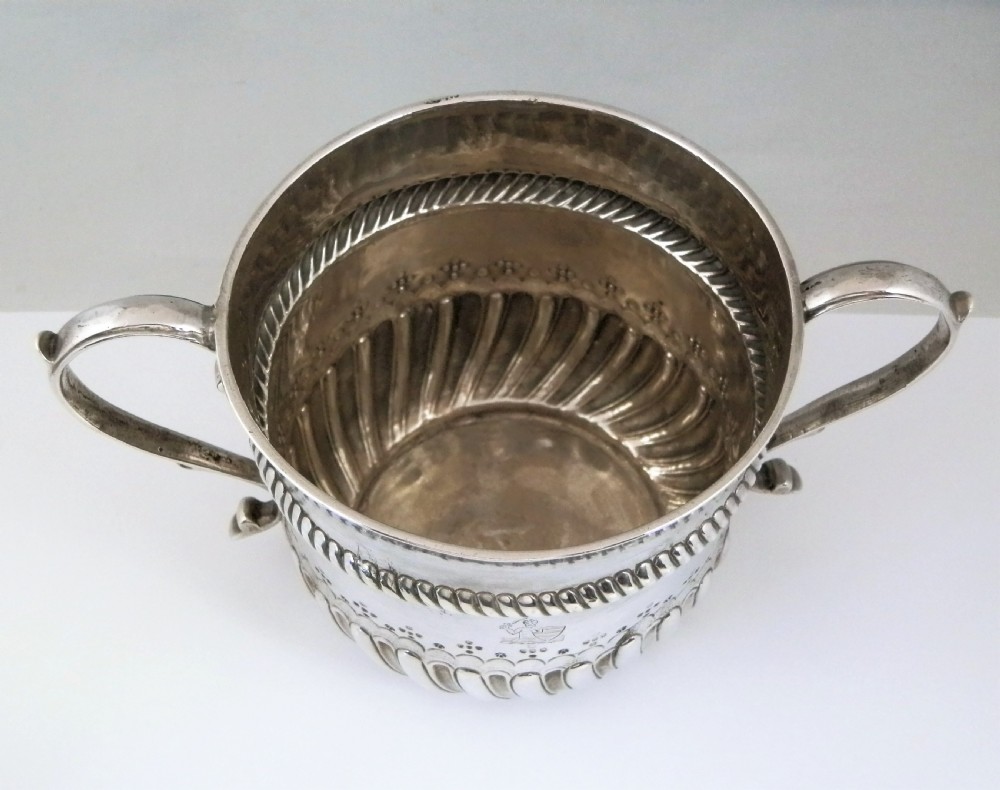 A 1694 William and Mary sterling silver porringer with twin scroll handles, rope-moulded girdle, lower spiral fluting, and an engraved armorial crest. View V