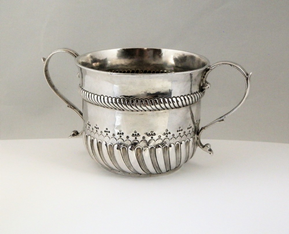 A 1694 William and Mary sterling silver porringer with twin scroll handles, rope-moulded girdle, lower spiral fluting, and an engraved armorial crest. View IX