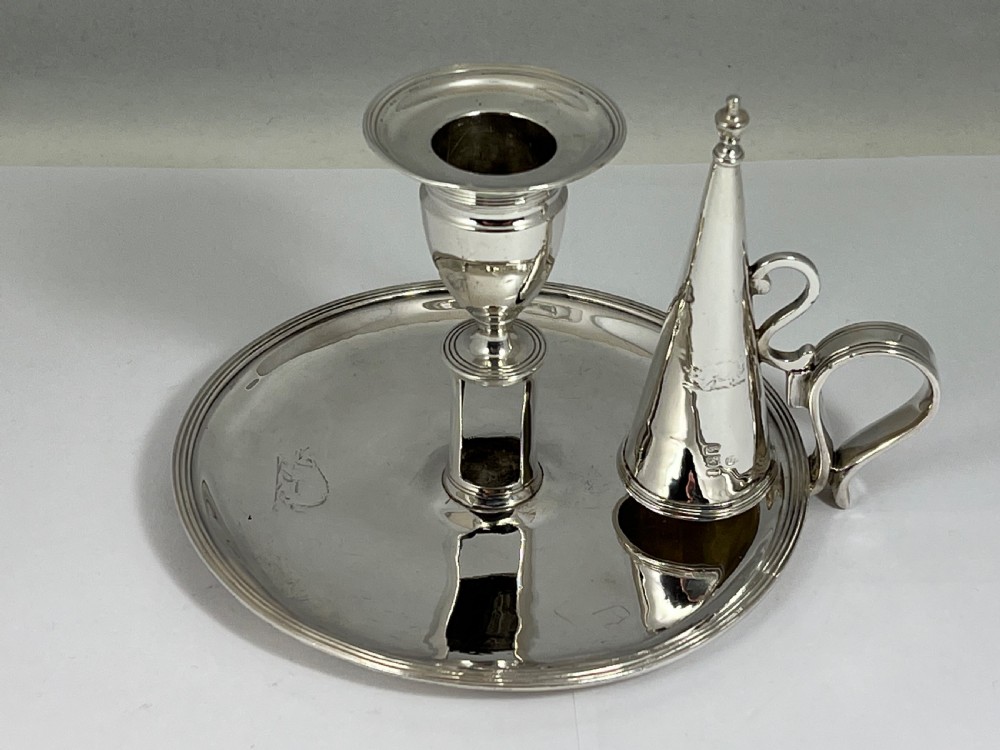 A George III sterling silver chamberstick featuring a circular base pan, a raised central candle socket, a curved side handle, and a removable conical snuffer.