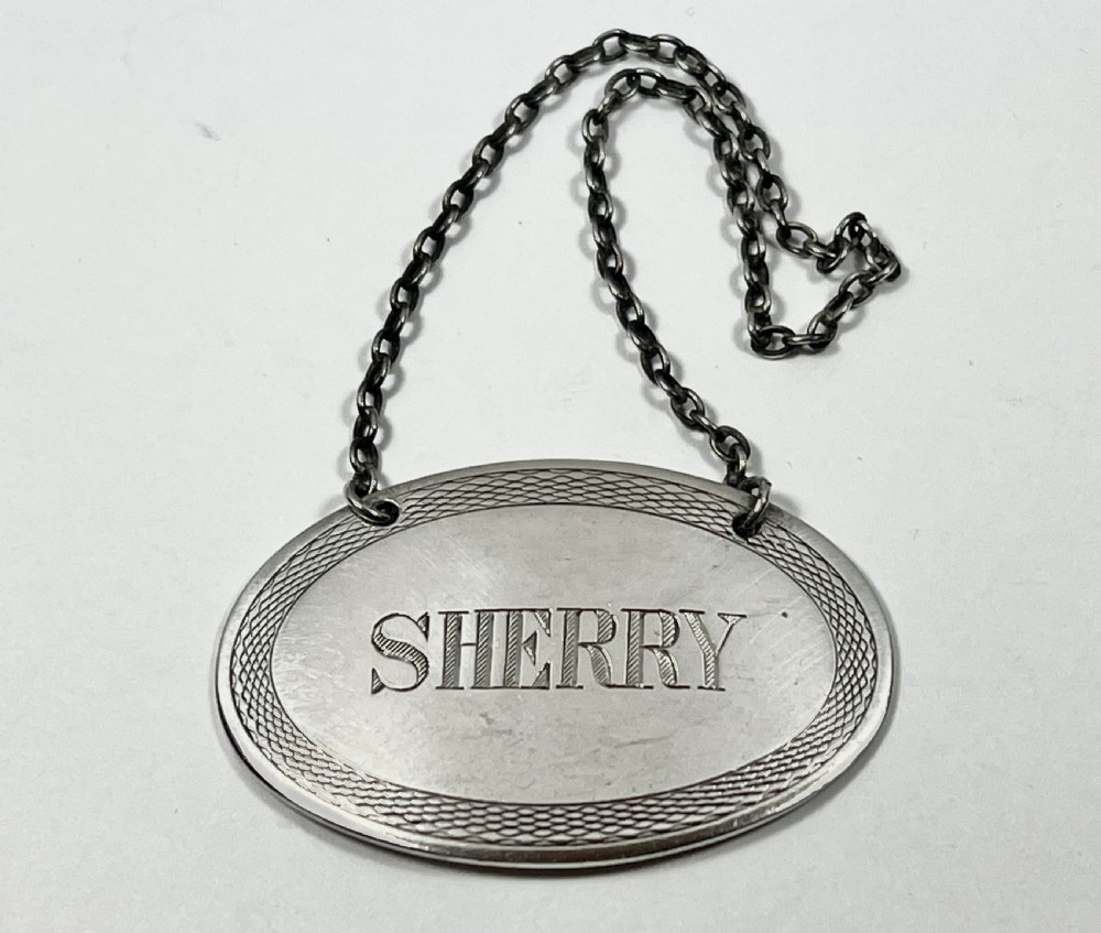 An oval sterling silver decanter label engraved with the word SHERRY and an engine-turned border pattern, suspended from a silver chain. View II