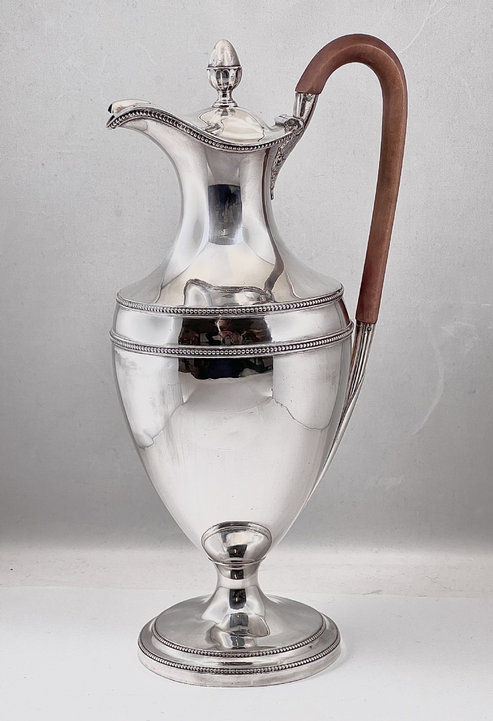 An urn-shaped sterling silver ewer with beaded border trim, a pinecone finial, and a high arched wooden handle.