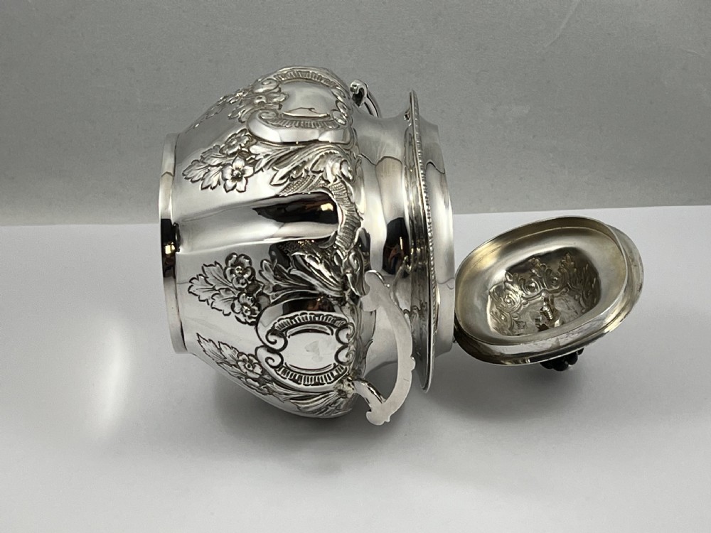 A sterling silver tea caddy featuring twin side handles, relief repousse floral scrollwork, and a domed lid topped with a dark fluted finial. View II