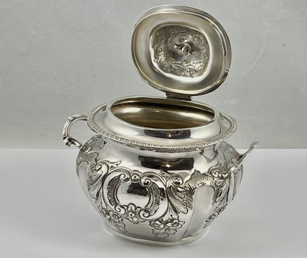 A sterling silver tea caddy featuring twin side handles, relief repousse floral scrollwork, and a domed lid topped with a dark fluted finial. View III