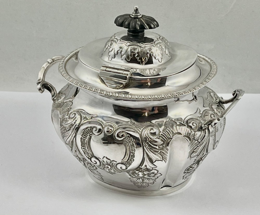 A sterling silver tea caddy featuring twin side handles, relief repousse floral scrollwork, and a domed lid topped with a dark fluted finial. View VI