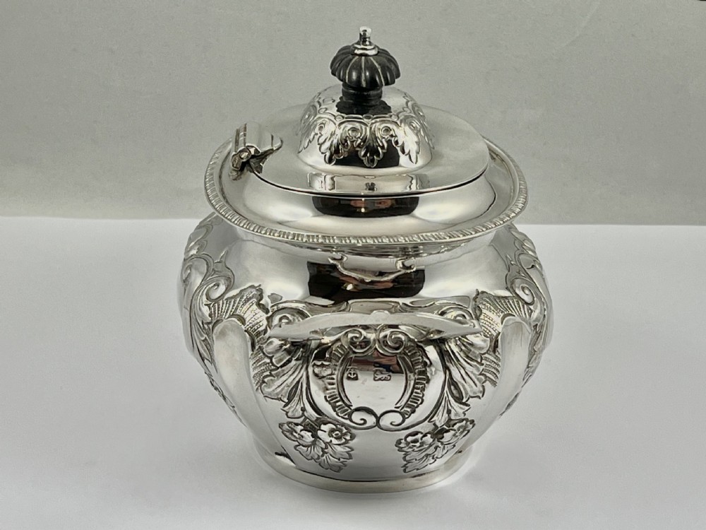 A sterling silver tea caddy featuring twin side handles, relief repousse floral scrollwork, and a domed lid topped with a dark fluted finial. View VII