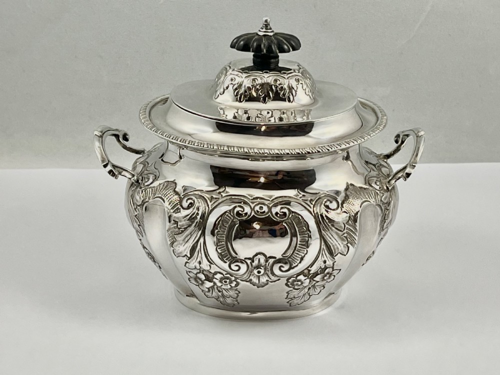 A sterling silver tea caddy featuring twin side handles, relief repousse floral scrollwork, and a domed lid topped with a dark fluted finial. View IX