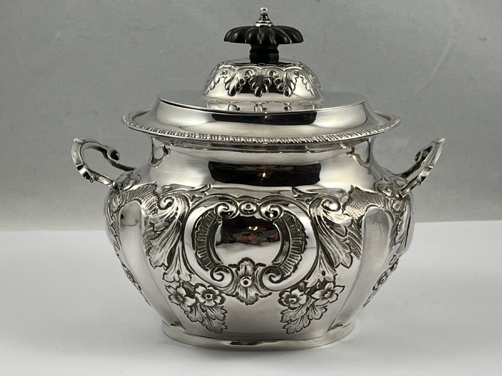 A sterling silver tea caddy featuring twin side handles, relief repousse floral scrollwork, and a domed lid topped with a dark fluted finial. View X