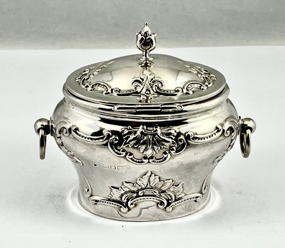An Edwardian sterling silver tea caddy featuring repousse scrollwork motifs, twin drop handles, and a fitted lid with a finial. View II