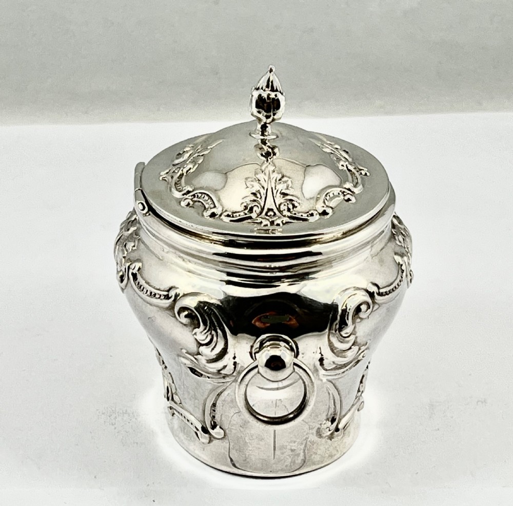An Edwardian sterling silver tea caddy featuring repousse scrollwork motifs, twin drop handles, and a fitted lid with a finial. View III