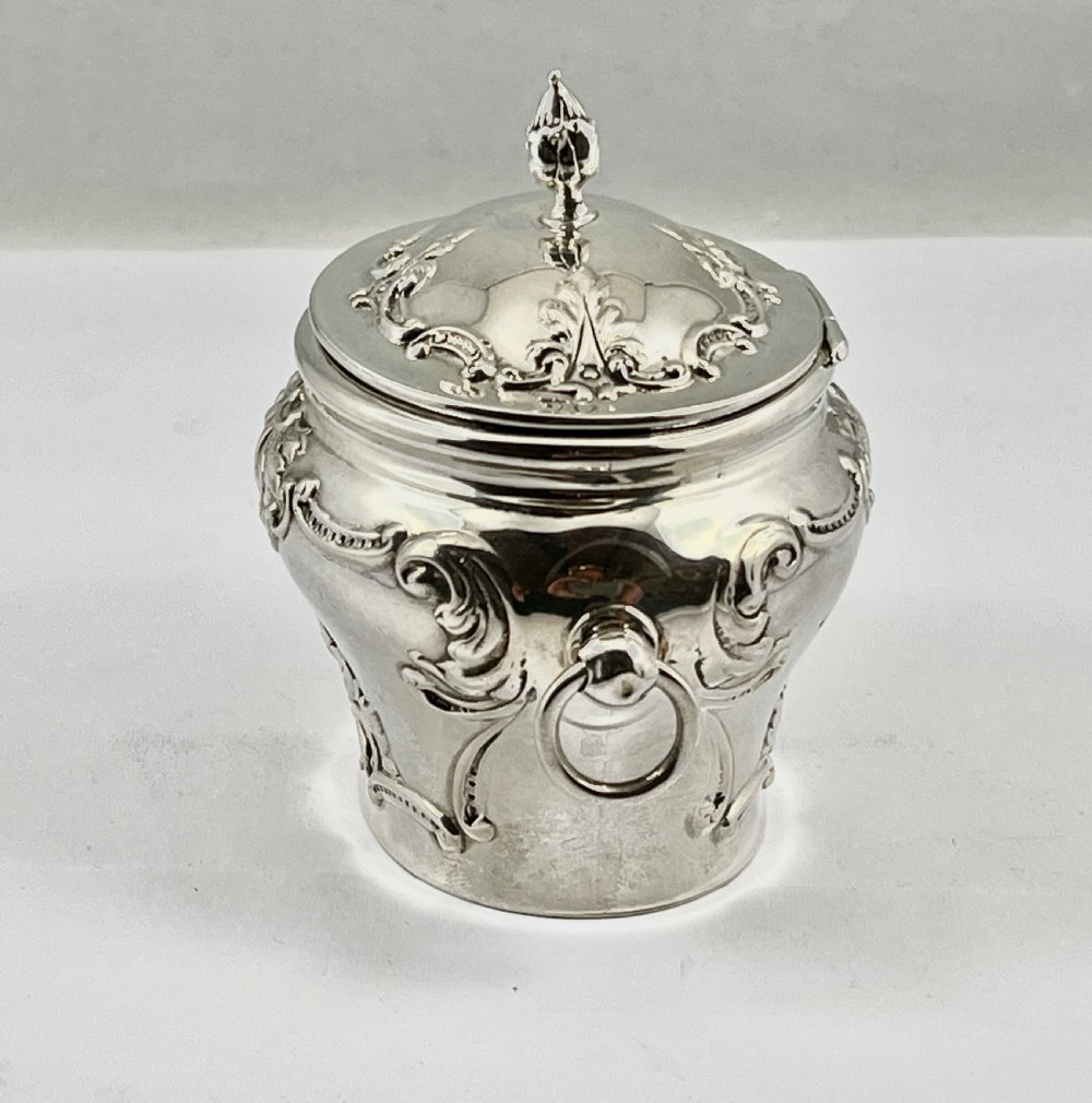 An Edwardian sterling silver tea caddy featuring repousse scrollwork motifs, twin drop handles, and a fitted lid with a finial. View V