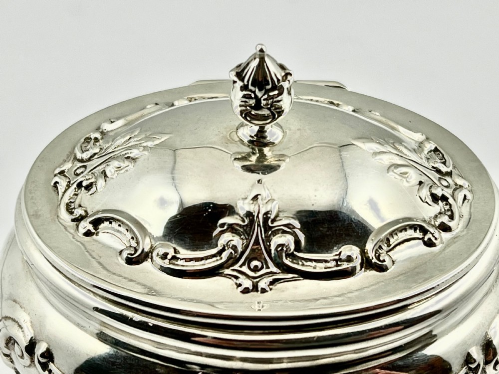 An Edwardian sterling silver tea caddy featuring repousse scrollwork motifs, twin drop handles, and a fitted lid with a finial. View VII