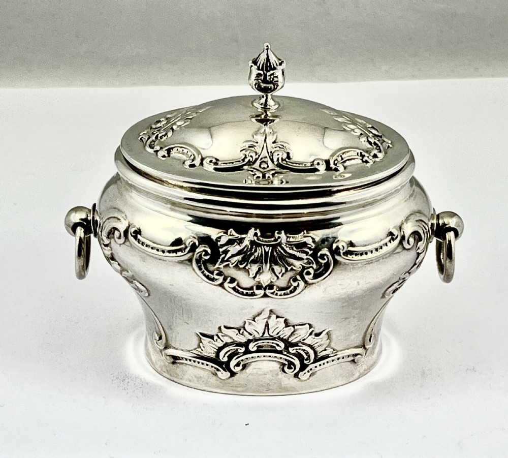 An Edwardian sterling silver tea caddy featuring repousse scrollwork motifs, twin drop handles, and a fitted lid with a finial. View VIII