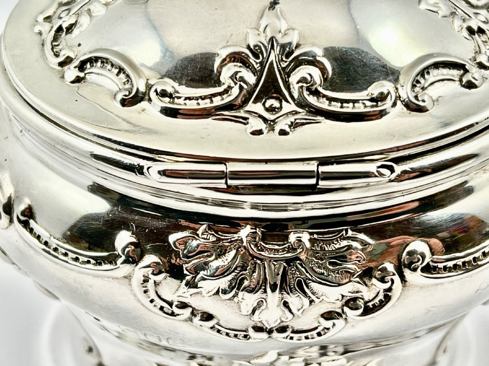 An Edwardian sterling silver tea caddy featuring repousse scrollwork motifs, twin drop handles, and a fitted lid with a finial. View X