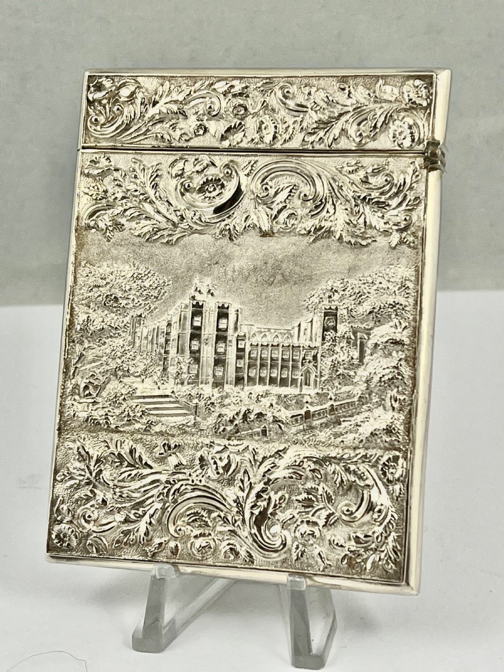 An Early Victorian sterling silver card case featuring detailed high-relief repousse carving of an architectural estate framed by scrollwork.
